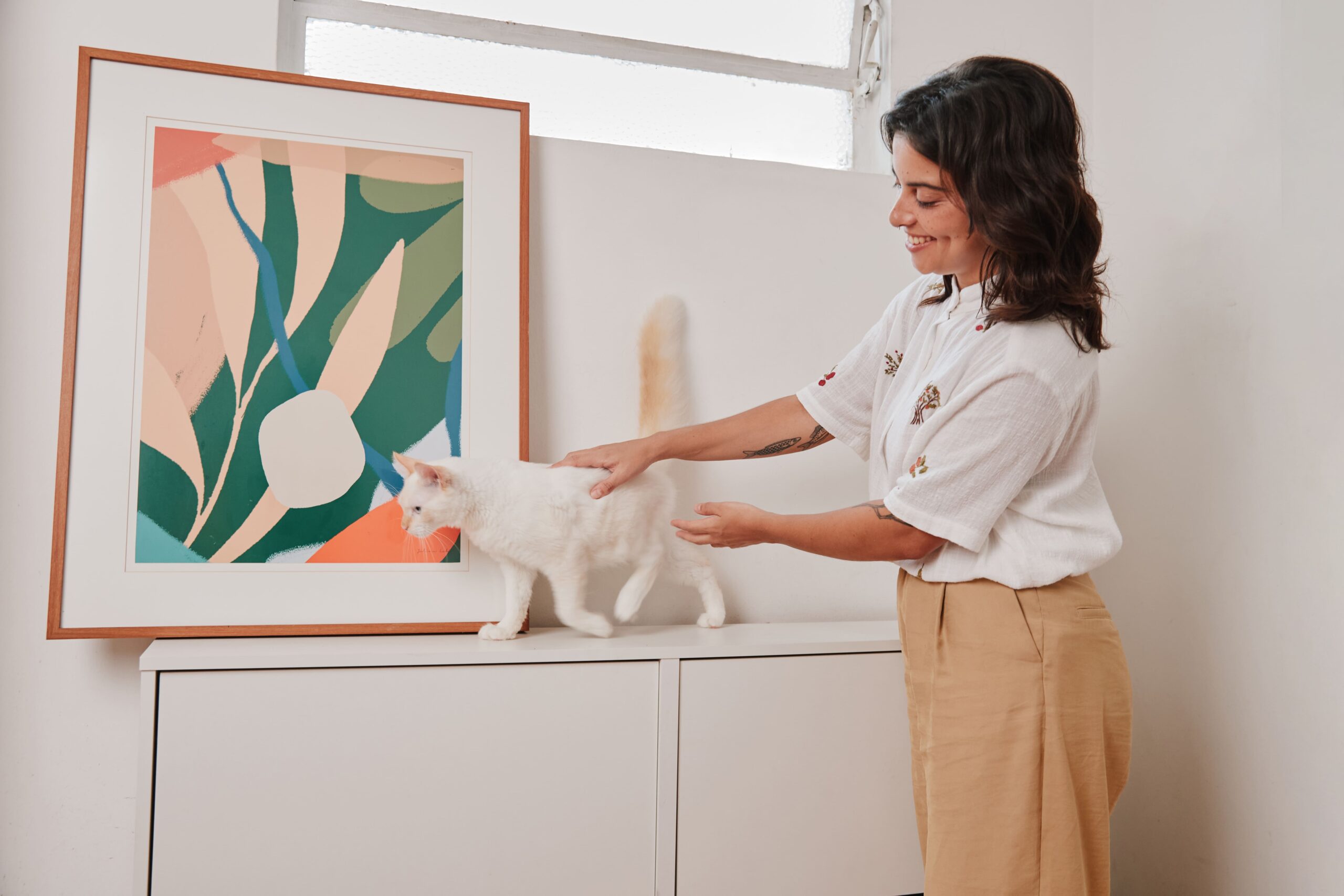 Mulher acariciando gato branco em cima de móvel com quadro.