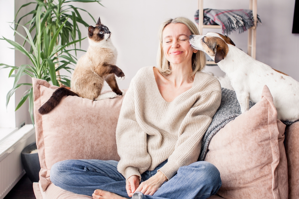 Mulher loira, com tricô bege e calça jeans, sentada no sofá entre um gato e um cachorro.