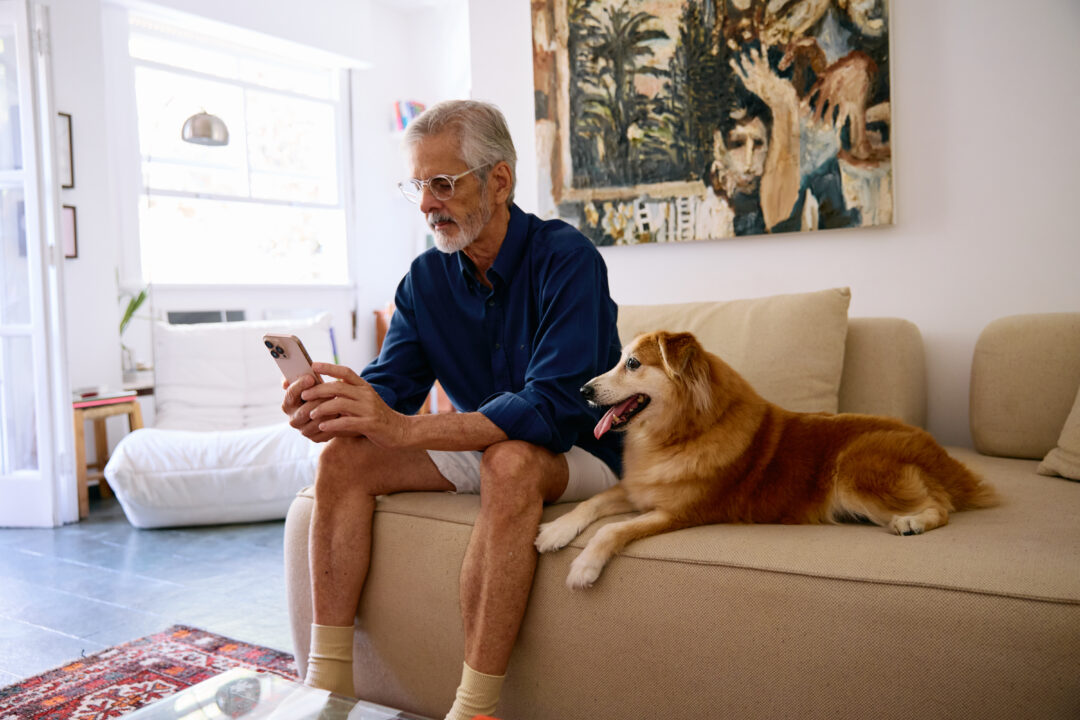Tutor olhando para o celular, sentado ao lado do cachorro no sofá.